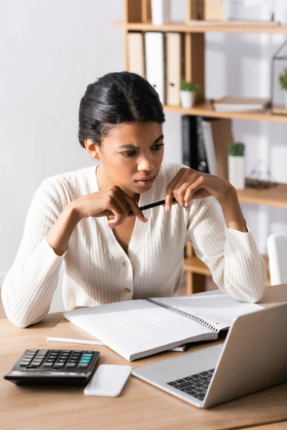 Задумчивая африканская американка смотрит на ноутбук, сидя за столом в офисе на размытом фоне - Фото, изображение