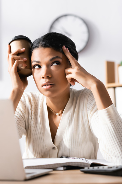 Tired african american woman looking away and holding cup of coffee near head in office on blurred foreground - Foto, imagen