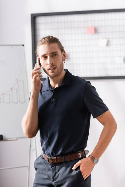 Confident businessman with hand in pocket looking away while talking on phone in office on blurred background - Photo, Image