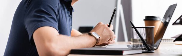 Vue recadrée de l'homme d'affaires avec stylo assis à la table de travail près d'un ordinateur portable sur fond flou, bannière - Photo, image