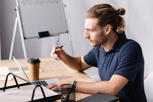 Zaostřený kancelářský pracovník s perem při pohledu na notebook při sezení na pracovním stole na rozmazaném pozadí - Fotografie, Obrázek
