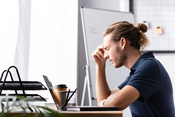 Gözü kapalı düşünceli bir ofis çalışanı laptopun yanındaki çalışma masasında arka planda bulanık bitkilerle oturuyor. - Fotoğraf, Görsel