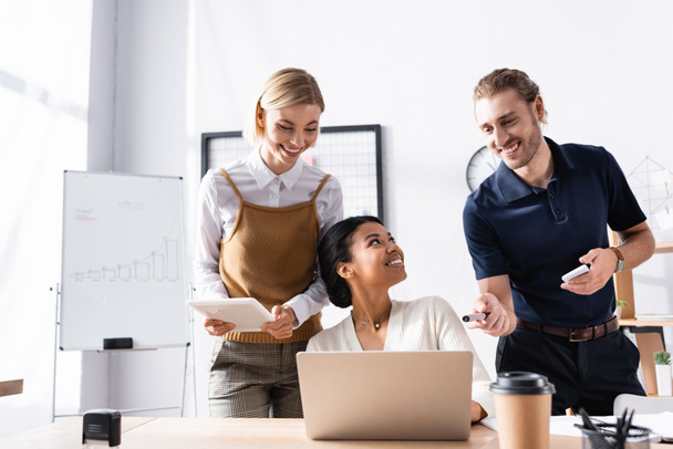 Szczęśliwi pracownicy biurowi stojący blisko afrykańskiej amerykanki siedzącej przy stole z laptopem w miejscu pracy - Zdjęcie, obraz