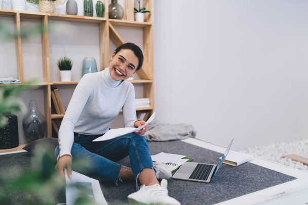 Optymistyczna freelancerka w nieformalnym ubraniu i okularach siedząca na podłodze i pracująca z dokumentami w domu podczas pracy na laptopie - Zdjęcie, obraz