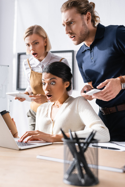 Шоковані багатокультурні офісні працівники дивляться на ноутбук на робочому місці з розмитим тримачем ручок на передньому плані
 - Фото, зображення