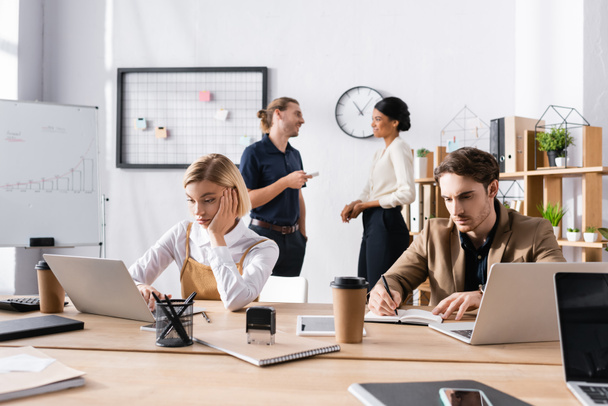 Скучные офисные работники используют ноутбук и пишут на ноутбуке, сидя за столом, с мультикультурными коллегами говорят на заднем плане - Фото, изображение