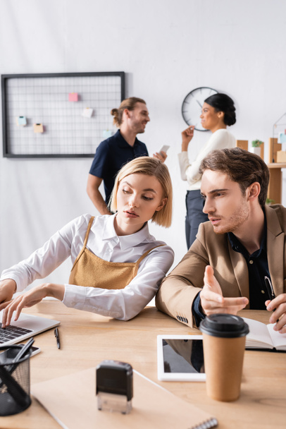 Konzentrierte blonde Frau tippt auf Laptop, während ein Mann in der Nähe am Arbeitsplatz sitzt, mit multikulturellen Büroangestellten auf verschwommenem Hintergrund - Foto, Bild