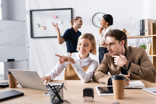 Орієнтований чоловік сидить на робочому місці біля жінки, вказуючи пальцем, використовуючи ноутбук з багатокультурними офісними працівниками на фоні
 - Фото, зображення