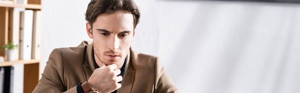 Thoughtful young adult man with hand near chin looking away in office on blurred background, banner - Fotoğraf, Görsel