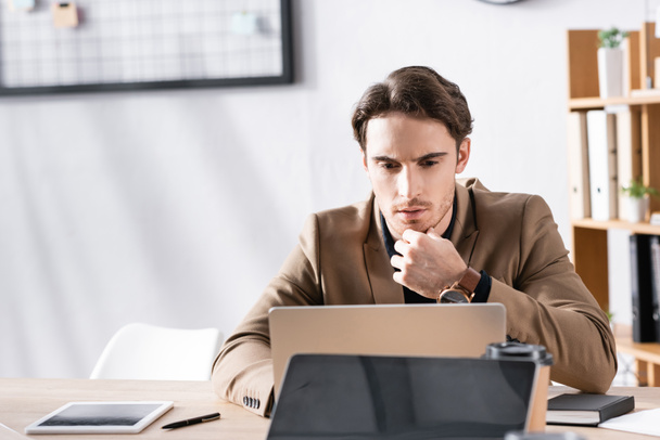 Thoughtful office worker looking at laptop while sitting at desk in office on blurred background - Photo, Image