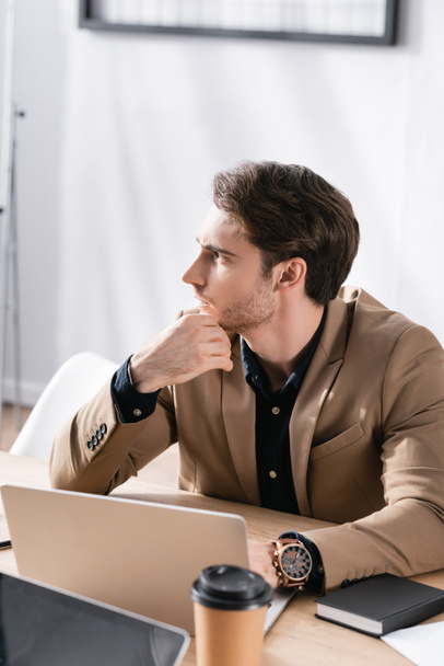 Thoughtful office worker looking away while sitting at workplace with laptop on blurred background - Photo, Image