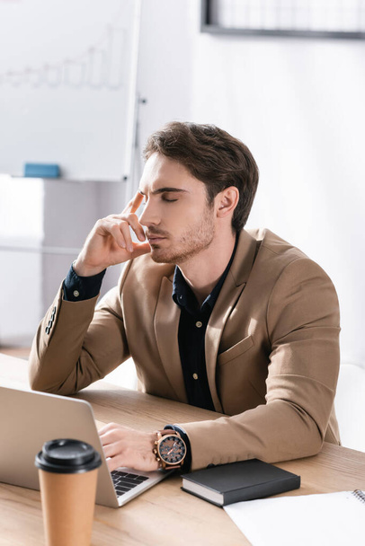 Продуманий бізнесмен із закритими очима тримає руку біля голови, сидячи за столом з ноутбуком на розмитому фоні
 - Фото, зображення