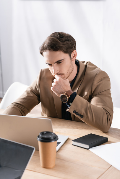 Вдумчивый взрослый мужчина в формальной одежде смотрит на ноутбук на столе в офисе - Фото, изображение