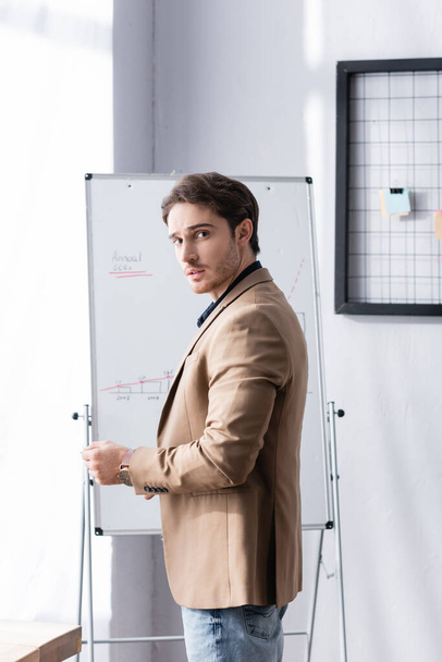 Důvěryhodný podnikatel se dívá do kamery, zatímco stojí poblíž flipchartu s grafikou v kanceláři - Fotografie, Obrázek