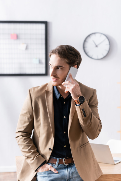 Mann mit der Hand in der Tasche, lächelnd beim Telefonieren, gestützt auf den Tisch am Arbeitsplatz vor verschwommenem Hintergrund - Foto, Bild