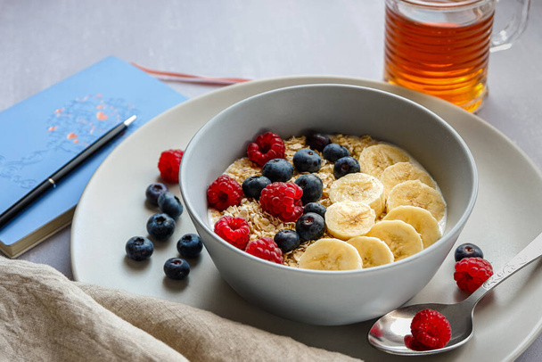 Боковой вид на здоровый завтрак с овсянкой в миске, банановыми ломтиками, малиной, черникой, чашкой чая и блокнотом с ручкой на светло-сером столе - Фото, изображение