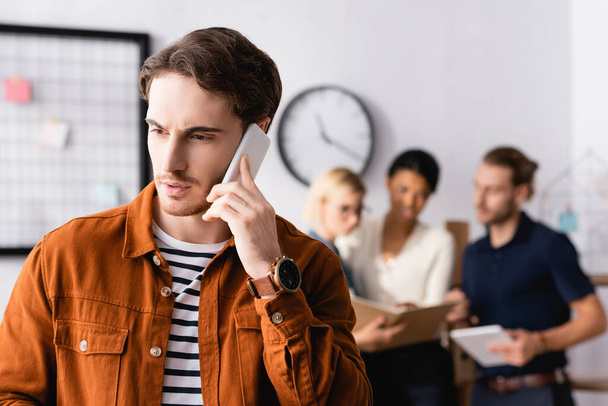 young businessman talking on smartphone near multiethnic businesspeople talking on blurred background - Фото, зображення