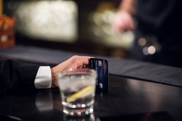 クレジットカードが閉じられている手の後ろに水とレモンのガラスがバーにあり - 写真・画像