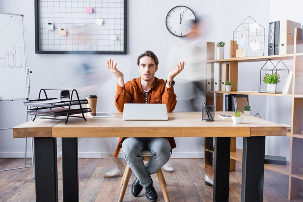 confused businessman showing shrug gesture while sitting at desk in open space office, motion blur - Photo, Image