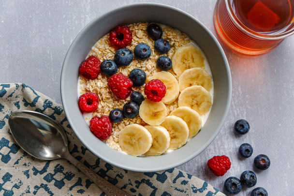 Vista superior de un desayuno saludable con harina de avena en un tazón, rodajas de plátano, frambuesa, arándanos y una taza de té en la mesa gris claro - Foto, imagen
