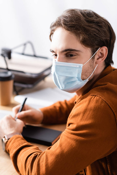 fiatal menedzser orvosi maszk ül a munkahelyen toll és notebook homályos háttér - Fotó, kép