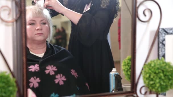 Een oudere vrouw met kort grijs haar doet styling in een schoonheidssalon. Een vrouw die voor een spiegel zit. Video - Video