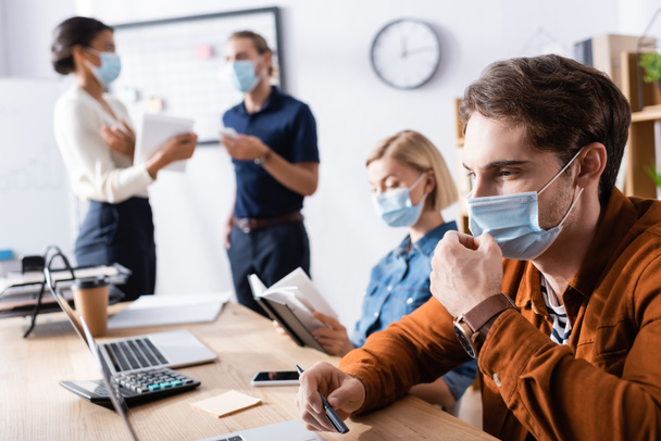 young businessman fixing medical mask on face while working near colleagues on blurred background - Foto, Bild