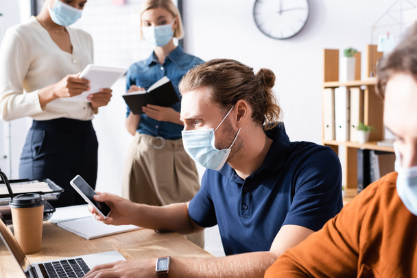 manager in medical mask using smartphone near multiethnic colleagues working on blurred background - 写真・画像