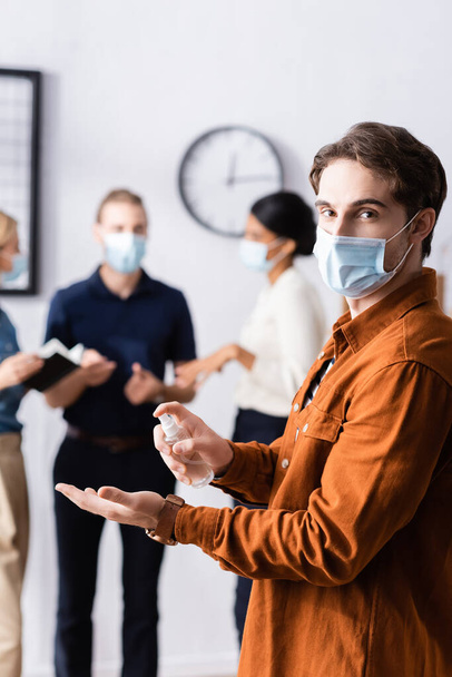 young businessman in medical mask holding sanitizer while looking at camera near colleagues on blurred background - Foto, imagen