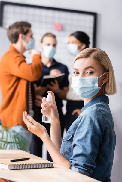szőke menedzser orvosi maszk gazdaság fertőtlenítő, míg a kollégák beszélnek homályos háttér - Fotó, kép