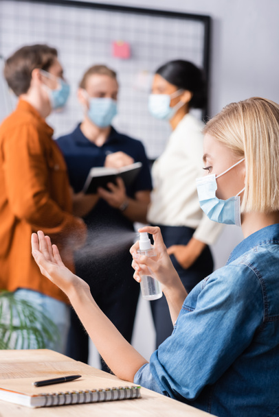 blonde businesswoman in medical mask spraying sanitizer near colleagues on blurred background - 写真・画像