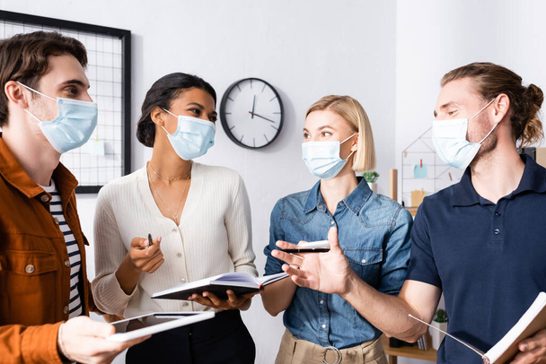 multiethnic businesspeople in medical masks holding notebooks and talking in office - Foto, Imagem