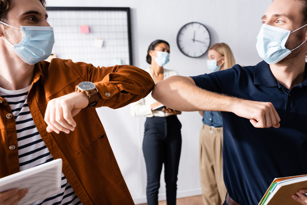 young managers in medical mask doing elbow bump gesture near businesswomen on blurred background - Photo, Image
