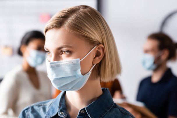 young, blonde manager in medical mask looking away near blurred multiethnic colleagues on background - Fotoğraf, Görsel