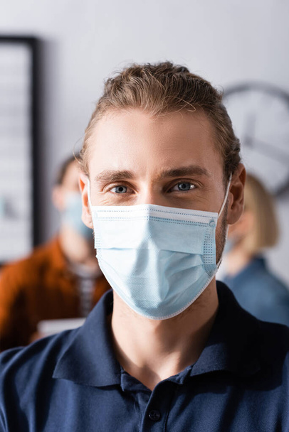 mladý manažer v lékařské masce při pohledu na kameru na rozmazaném pozadí - Fotografie, Obrázek
