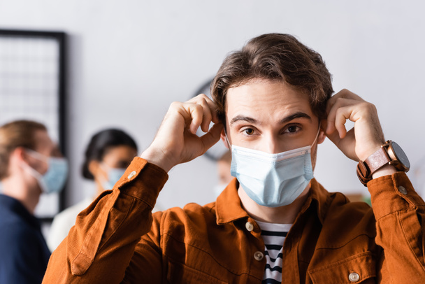 mladý manažer nasazení lékařské masky při pohledu na kameru v blízkosti rozmazané multikulturní kolegy na pozadí - Fotografie, Obrázek