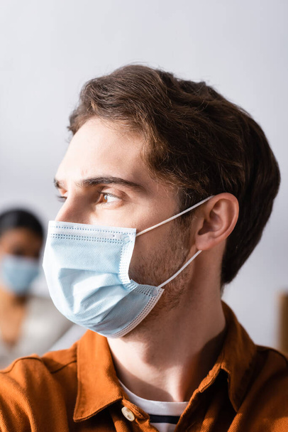 young businessman in medical mask looking away in office on blurred background - Foto, afbeelding