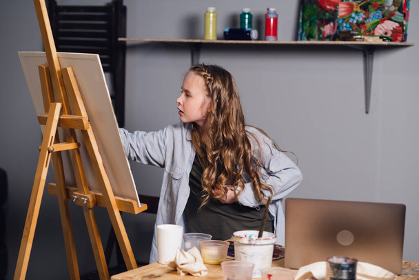 χαριτωμένο κορίτσι καλλιτέχνης αντλεί ελαιογραφία της από μια αναφορά από ένα φορητό υπολογιστή. - Φωτογραφία, εικόνα
