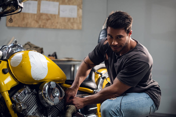 A young sports motorcycle repairman in a garage or workshop is working on a yellow bike. - Zdjęcie, obraz