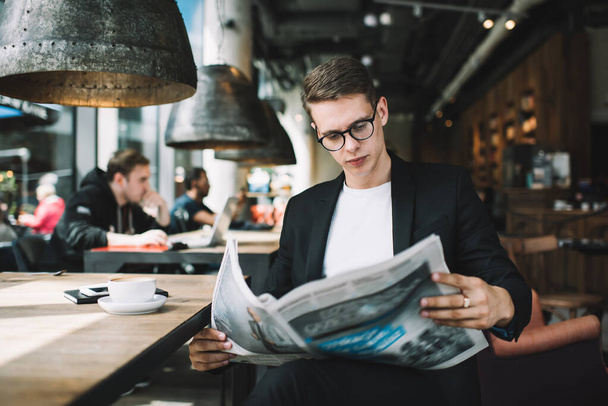 Concentrated elegant dressed man in eyeglasses attentively reading newspaper while sitting with cup of hot drink in dark trendy cafe - Foto, Bild