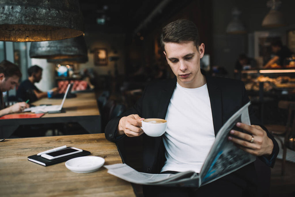 Concentrated young well dressed businessman reading newspaper and drinking hot drink while sitting with cellphone and notepad in stylish cafe - Foto, Imagen