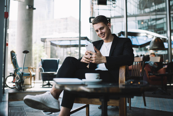 Happy male in casual clothes smiling and looking at smartphone while sitting at table with cup of coffee resting while break - 写真・画像