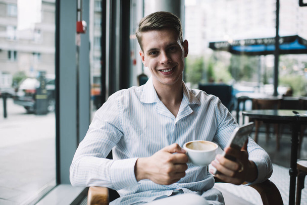 Positive young man sitting on chair in modern cafe drinking coffee and surfing phone while smiling and looking at camera in daylight - Fotografie, Obrázek