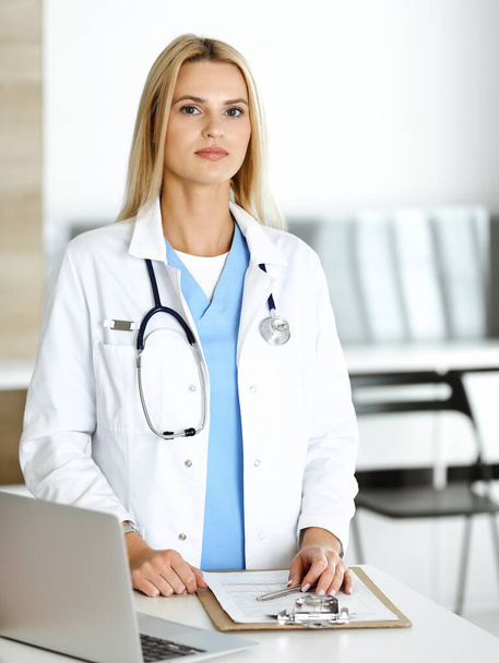 Doctor woman at work in hospital excited and happy of her profession. Blonde physician controls medication history records and exam results while using clipboard. Medicine and healthcare concept - Foto, Imagem