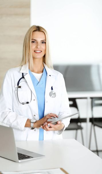 Ärztin bei der Arbeit in der Klinik aufgeregt und glücklich über ihren Beruf. Die blonde Ärztin lächelt, während sie den Tablet-Computer bedient. Medizinkonzept - Foto, Bild