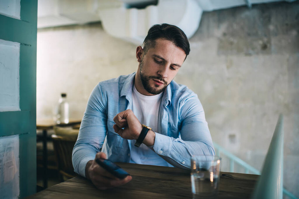 Serious bearded man in casual clothes sitting in chair in modern cafe with smartphone and glass of water and checking time on wristwatch - Фото, изображение