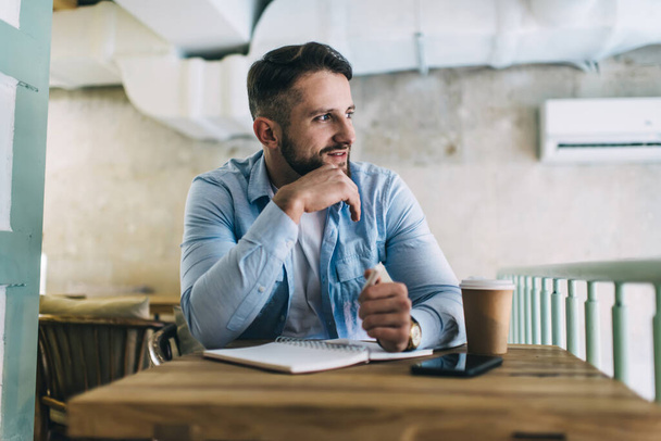Inspired handsome bearded man in casual shirt working at table in cafe with notebook and cup of coffee while looking away - Foto, Imagen