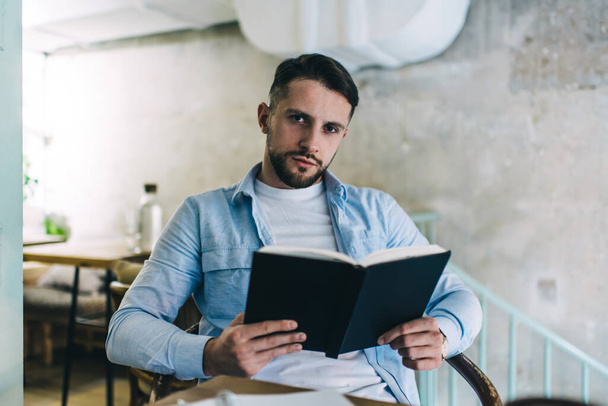 Young male with beard in casual clothes reading book while sitting in chair at table in well lit cafe and looking at camera - Фото, изображение