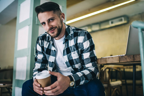From below of glad stylish man in checkered shirt sitting in creative cafe with hot beverage to go and listening to songs in wireless earphones while enjoying weekend - 写真・画像