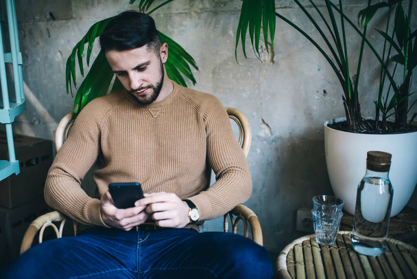 Serious bearded man wearing in sweater with cellphone resting in wooden armchair before drinking water from glass bottle in modern cafe - Foto, Imagem